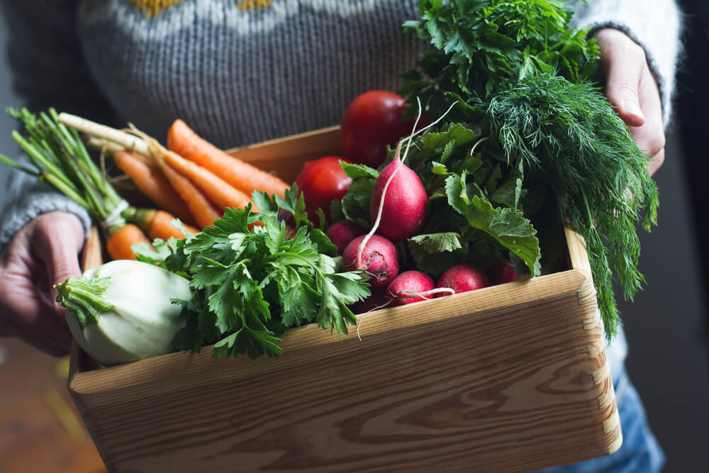 A gardener holding a large wooden crate stuffed with yummy veggies.