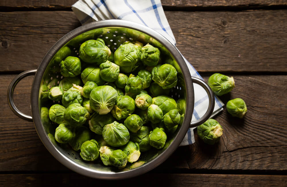 Fresh Brussels Sprouts sitting in a strainer atop a wooden table.