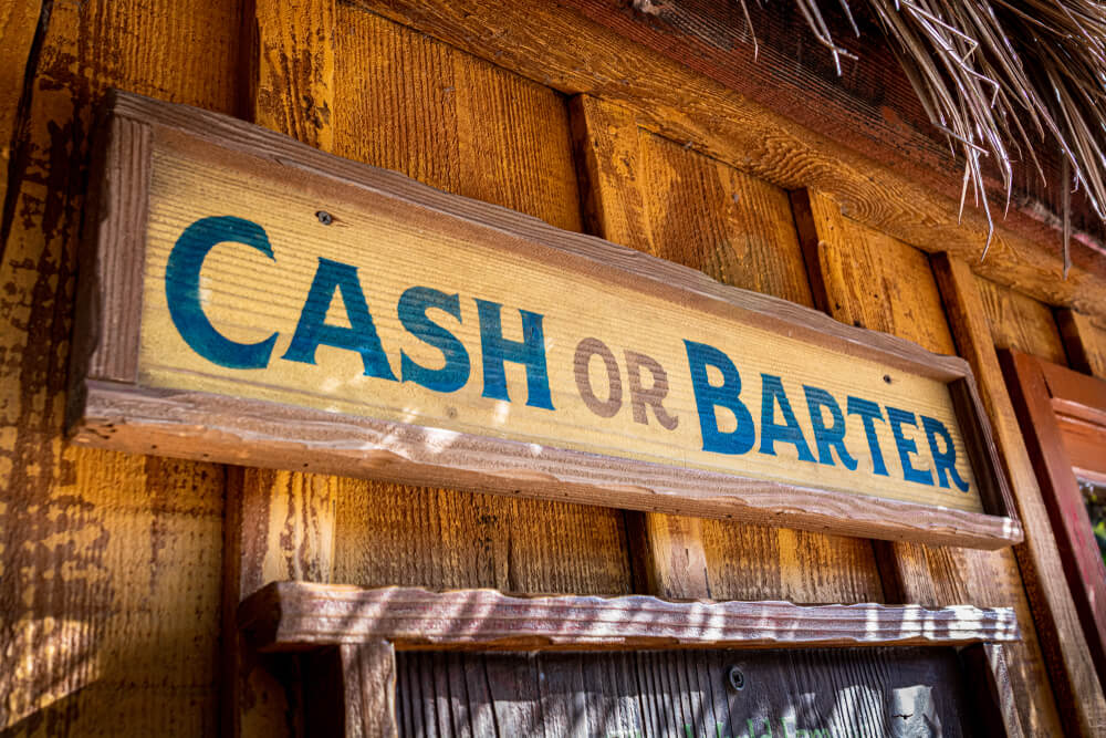A farmyard or rural store front banner sign promoting that they accept cash and barter.