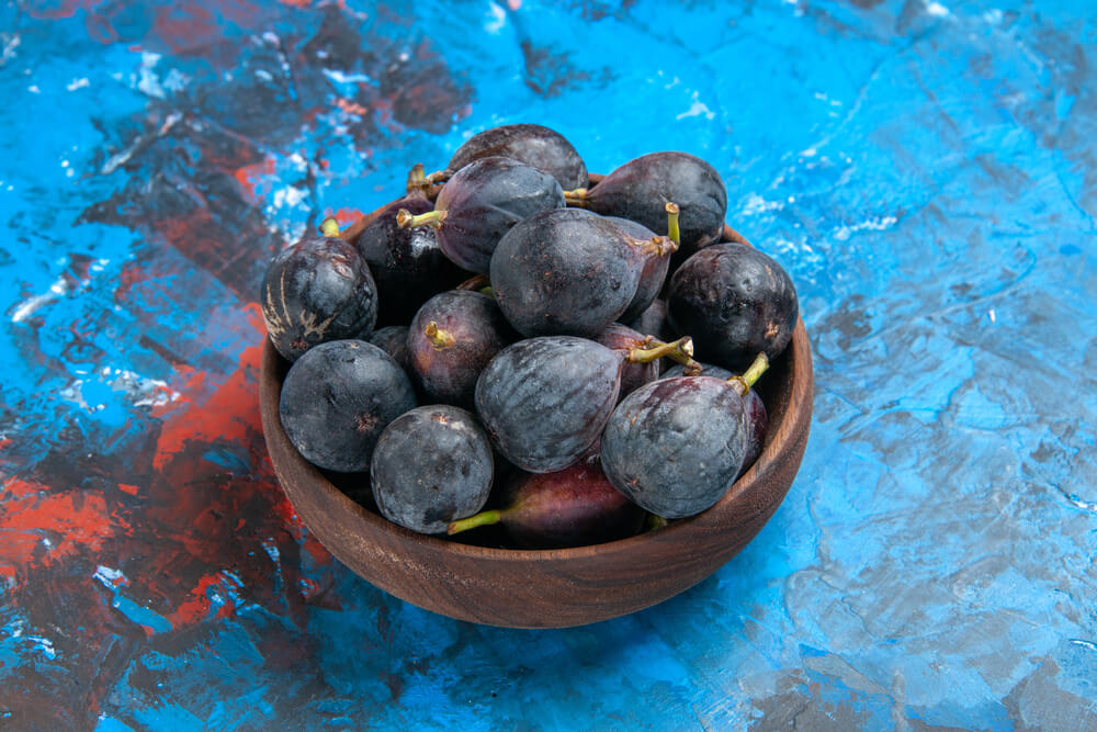 Black mission figs harvested in a brown pot atop a blue table.