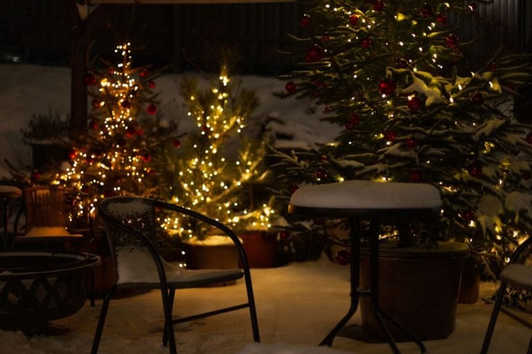 Beautiful winter view with Christmas trees, snow, lights, chair, and table on patio at night.